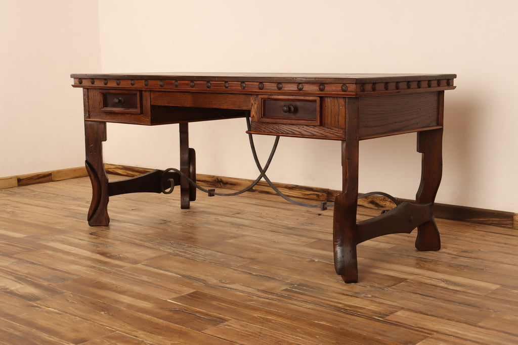 Wooden desk with three drawers on a wooden floor against a beige wall, decorative nailhead trim.