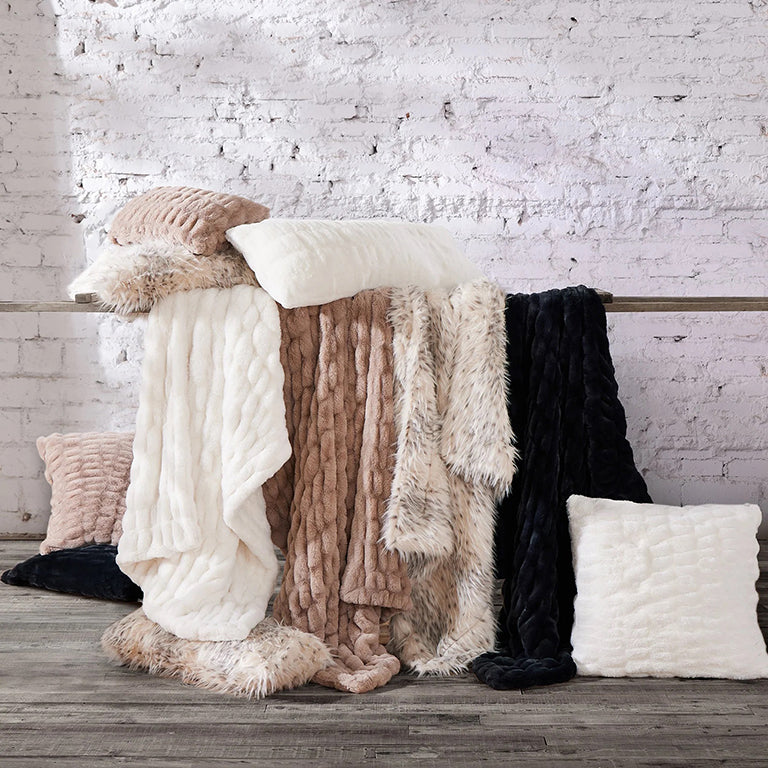 Collection of soft textured blankets and pillows on a wooden surface with a white brick wall background.