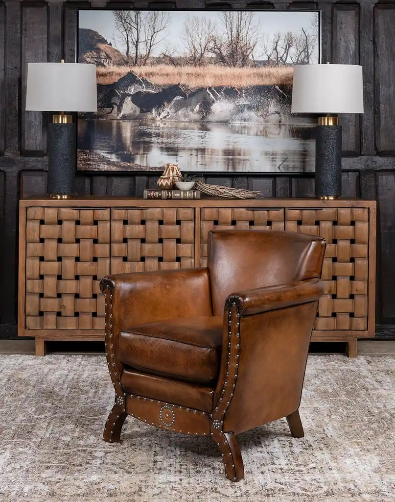 Brown leather armchair in front of a woven wooden console table with a large landscape painting above.