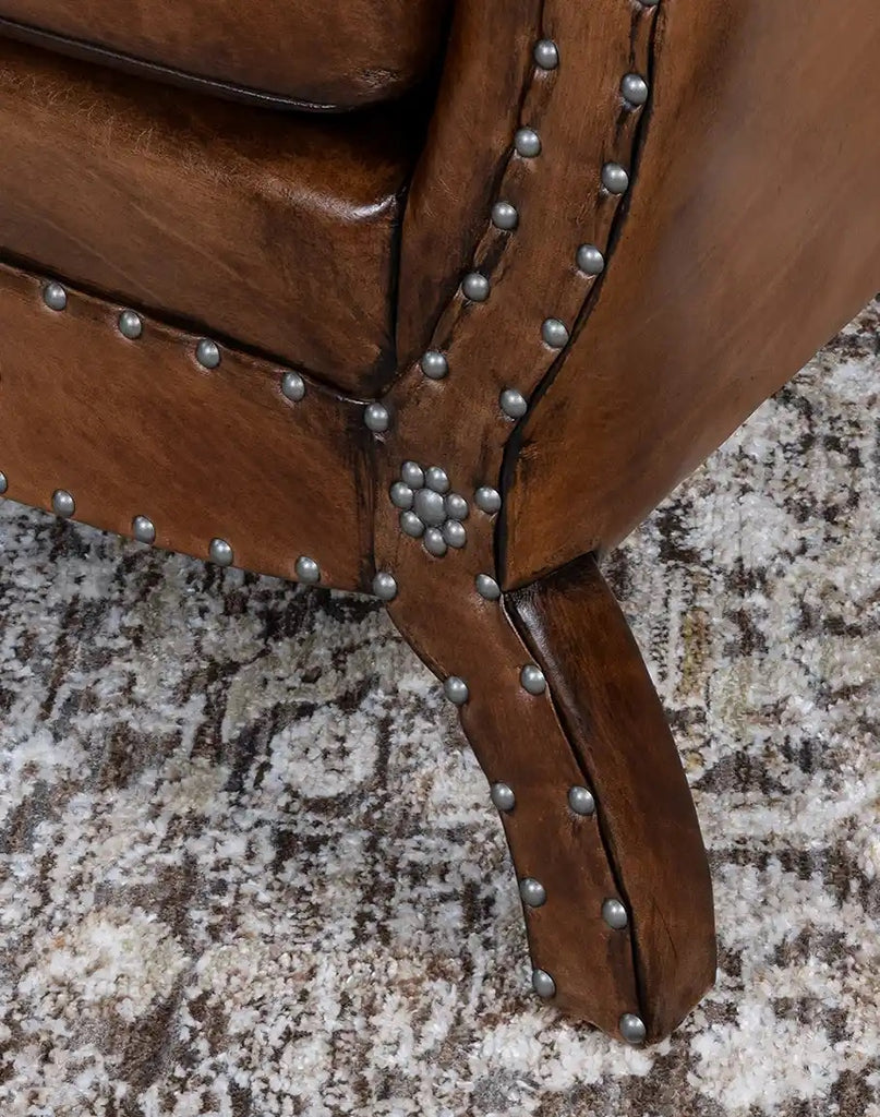 Close-up of a brown leather armchair with metal studs on a textured carpet.