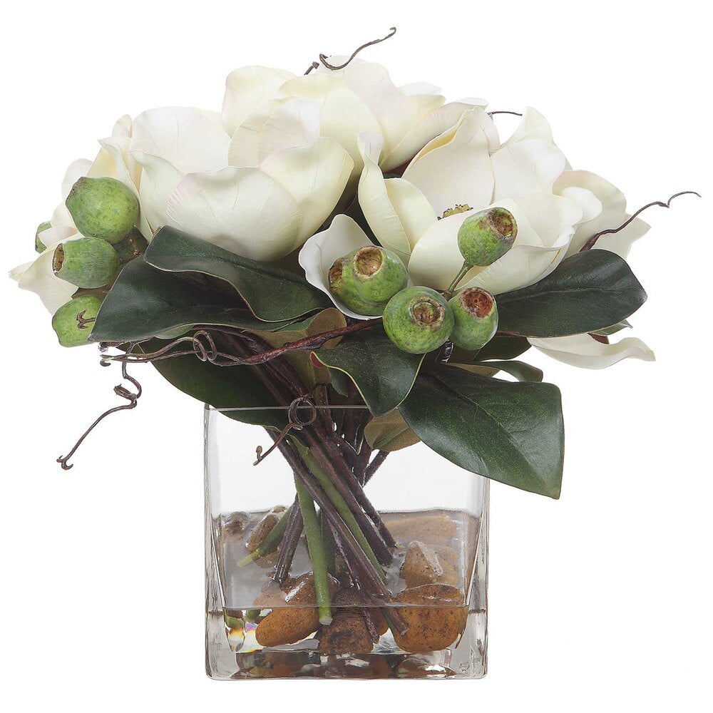 A timeless white magnolia floral arrangement with accompanying seed pods, river stones, and faux water in a transparent square glass vase.
