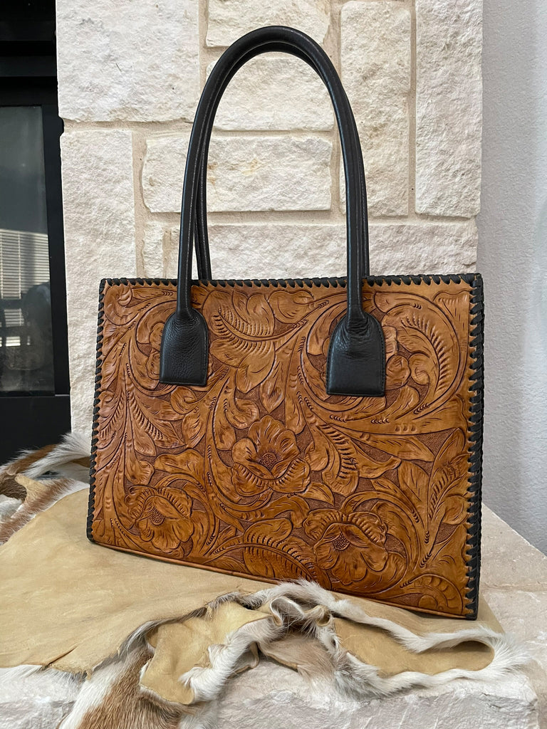 Brown leather handbag with dark brown handles on a textured surface with stone wall background