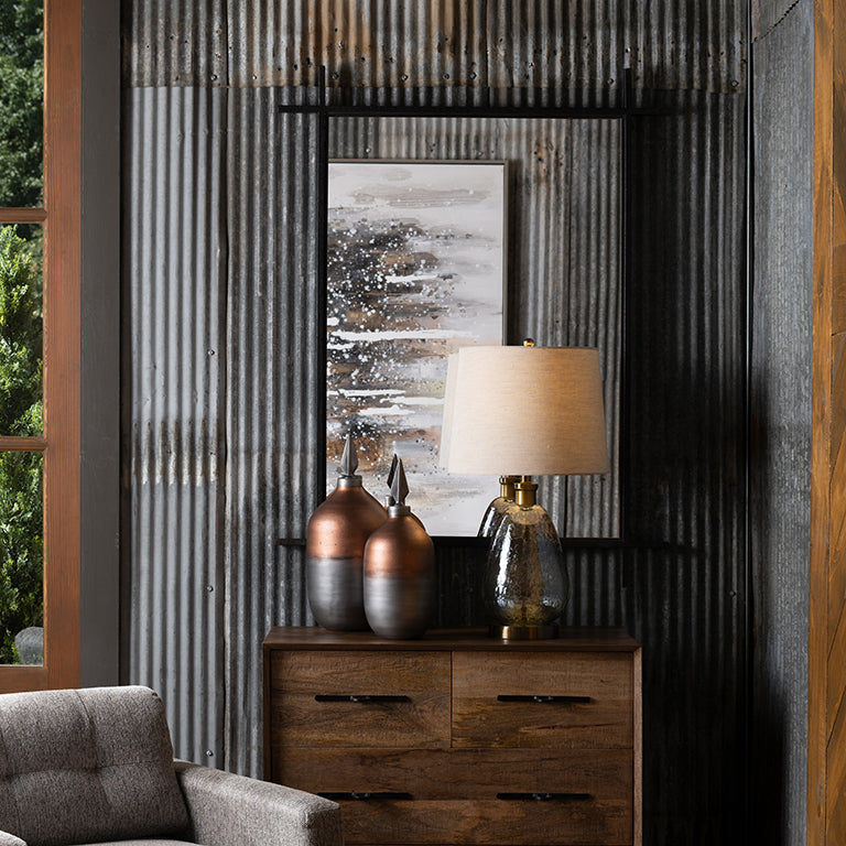 Decorative setting with a wooden dresser, lamp, and vases against a corrugated metal wall with mirror. Mirror has reflection of painting on opposite wall.