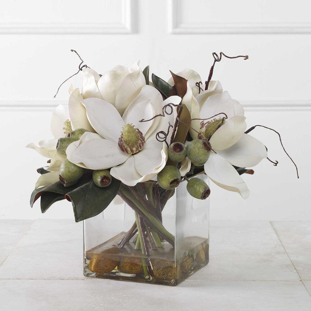 A charming centerpiece featuring white magnolia blossoms, seed pods, and river stones in a clear glass vase with faux water.