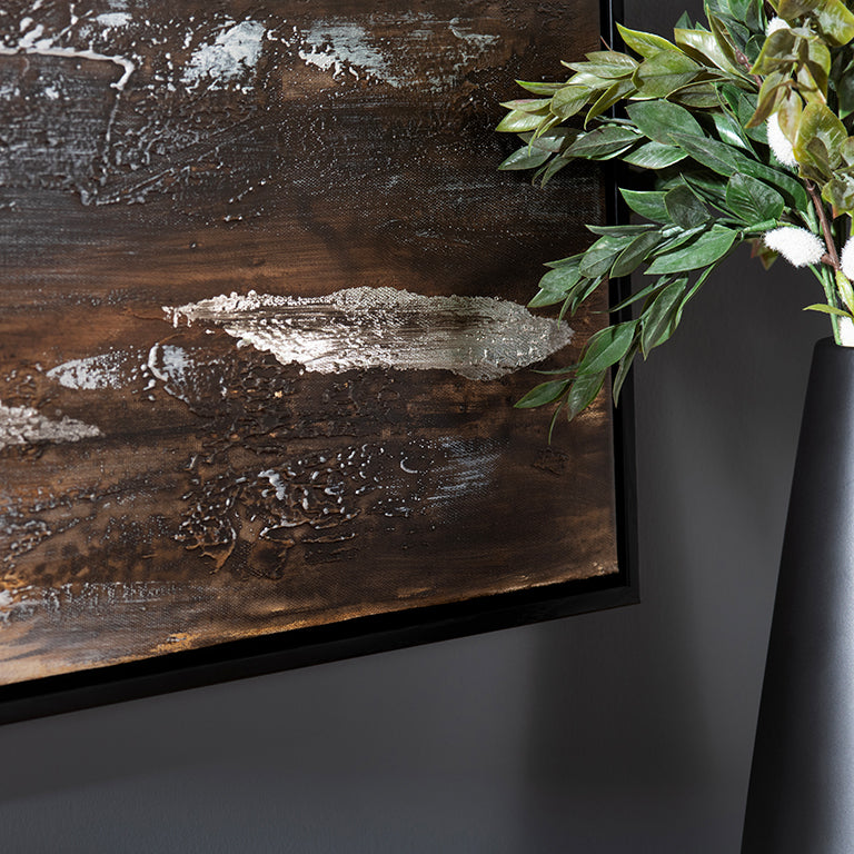 Wooden wall art with a textured finish next to a black vase with green leaves on a dark background