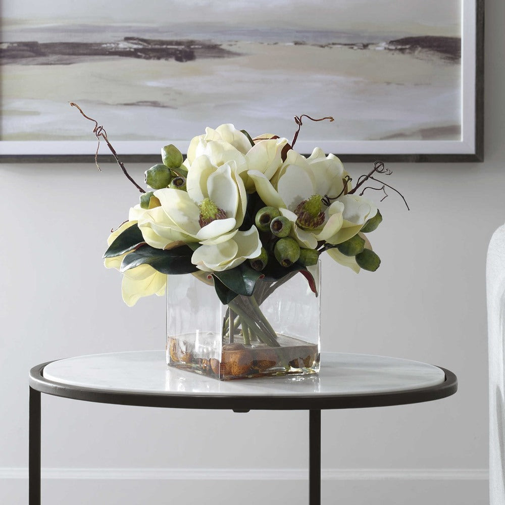 An arrangement of white magnolia blossoms and seed pods in a glass vase filled with river stones and faux water for added sophistication.