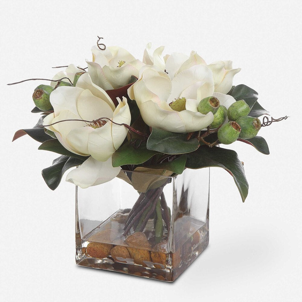 A stunning white magnolia bouquet featuring seed pods, natural river stones, and faux water arranged in a square glass vase.