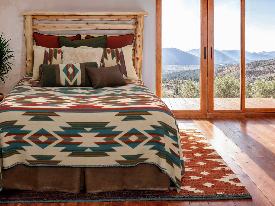 Wooded River Puesta Del Sol Value Bedding Set displayed on a bed, featuring vibrant Spice, Sage, Crème, and Camel colors with red and blue accents. Includes bed cover, two pillow shams, two euro shams, and one alt euro sham. Decorative stitching, faux fur, and leather fringe accents add rustic charm. Part of the 2025 Signature Washable Wool collection.